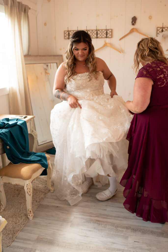 Bride putting on her left shoe while holding up her dress. Mother of the bride is helping 