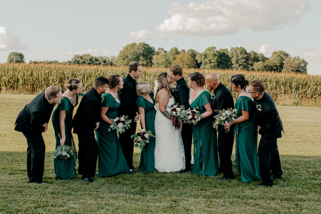 Full bridal party after ceremony. Bride and groom are kissing. Their party is looking in towards them. 