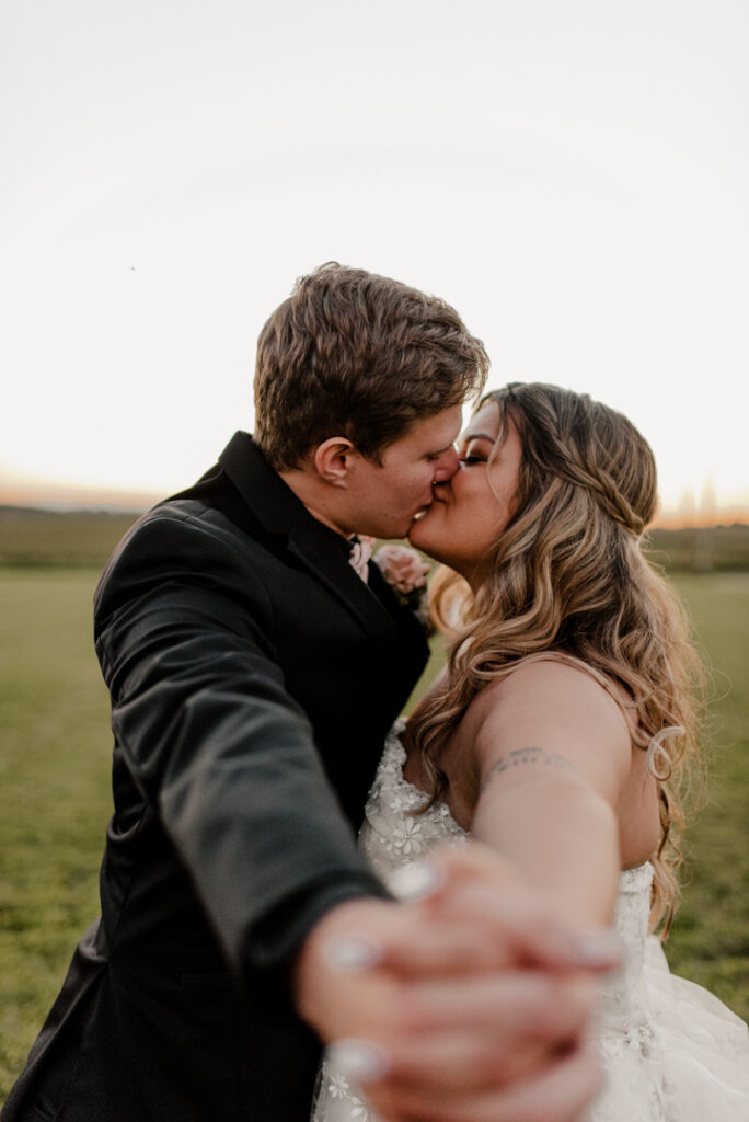 Bride and groom are kissing. Holding hands in front of the camera. The sun is setting behind them. 