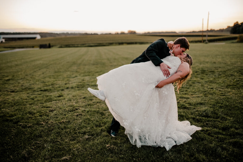 Groom is kissing bride while bending her backwards in a dip. The sun is setting behind them in a field. 