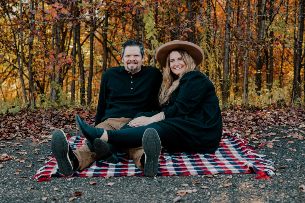 Man and woman sitting on a blanket on the ground. Both are smiling at the camera 