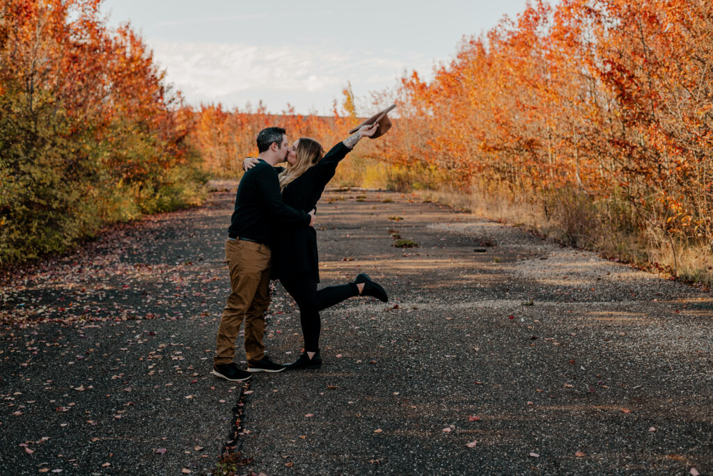 Wide photo of man and woman kissing. Her foot is kicked out and her hat is being thrown in the air. 