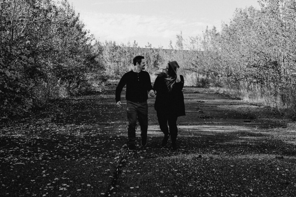 Black and white photo. Man and woman are running towards me while holding hands. She is holding onto her hat. 